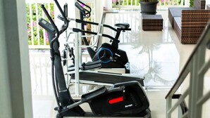 Un gimnasio en casa con equipo de ejercicio, incluyendo una caminadora y una elíptica, en un balcón con vistas a un jardín.