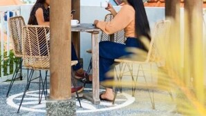 Un área de estar al aire libre techada con dos personas sentadas en una mesa, rodeadas de sillas de mimbre, y vistas de edificios al fondo.