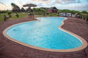 A swimming pool with a brick deck, surrounded by thatched-roof huts and greenery. - HOTEL HI MANANJARY (Mananjary)