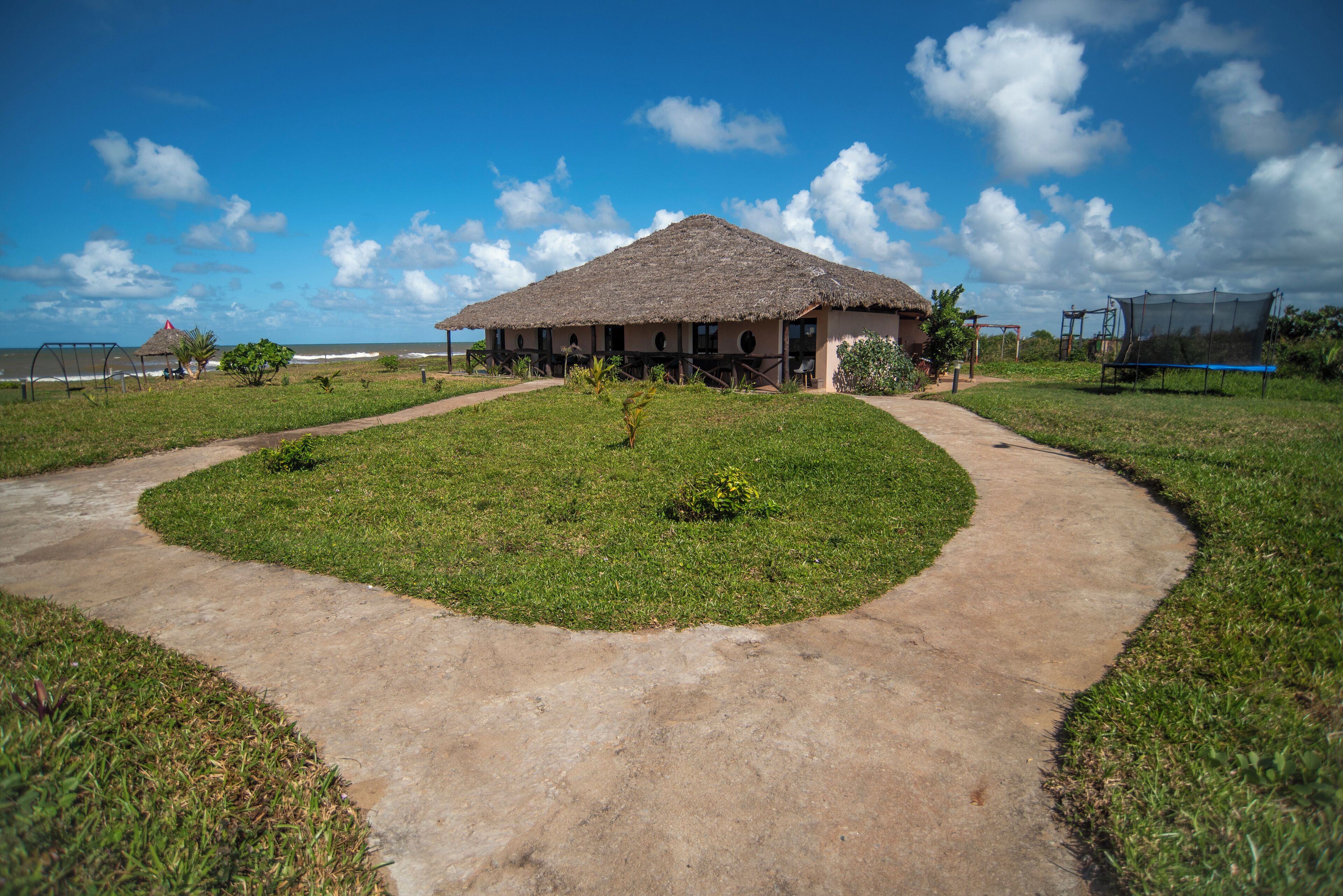 A thatched-roof building with a paved pathway leading to it, surrounded by grass and a few small plants.