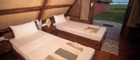A room with two beds, wooden beams, a thatched roof, and a view of the ocean.