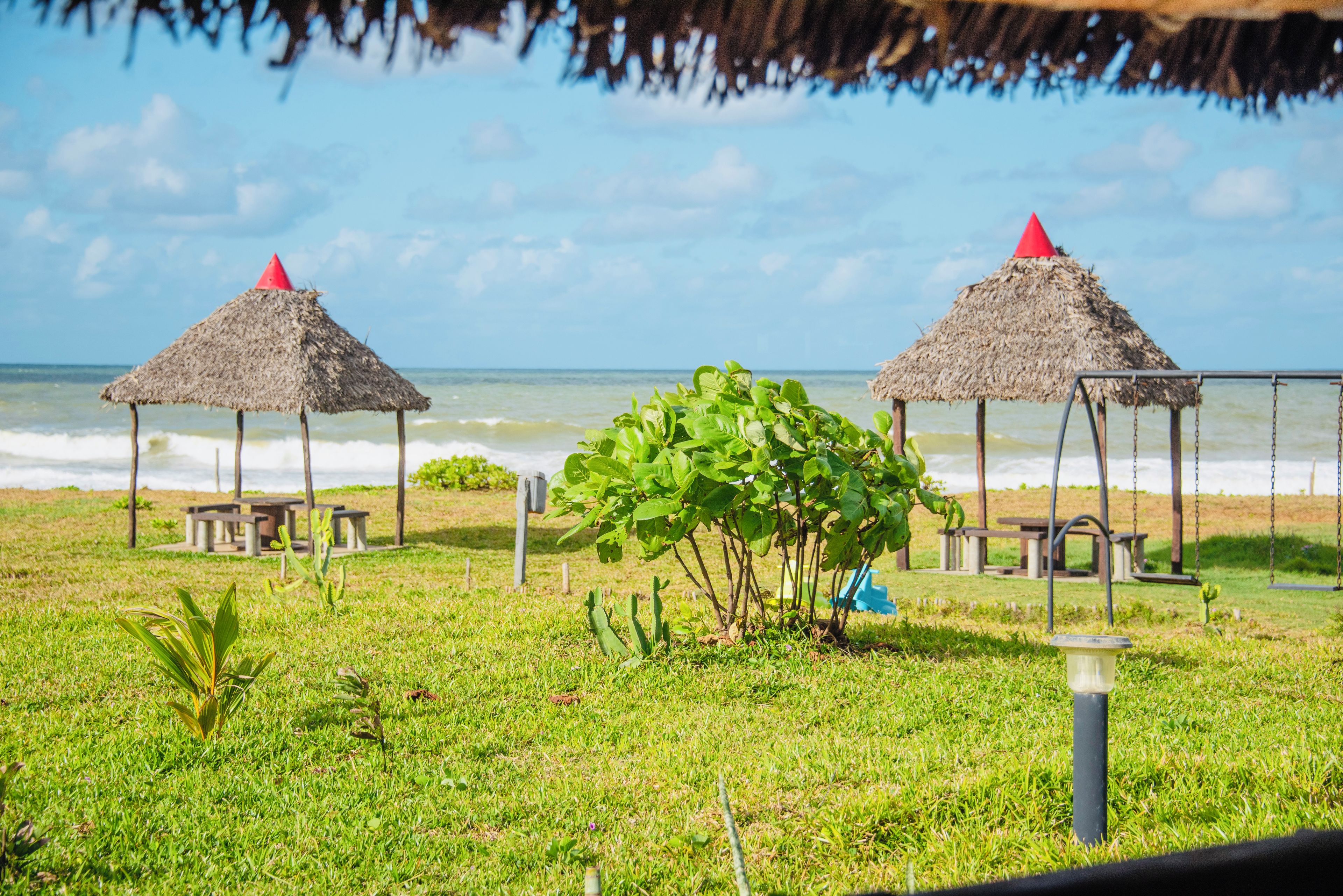 Uma praia com duas construções de palha, uma área gramada e o oceano ao fundo.