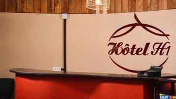 Un mostrador de recepción de hotel con un logotipo, una lámpara colgante y un cartel con un mensaje.