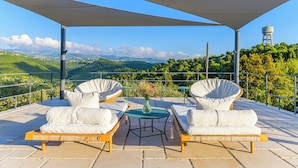 Une terrasse meublée de mobilier en osier, une table agrémentée d’un vase, et une vue sur des collines verdoyantes et un château d’eau.