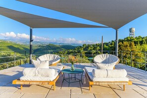 A terrace with wicker furniture, a table with a vase, and a view of green hills and a water tower. - Amani Luxury 4BR Villa & Pool in Batroun (Batroun)