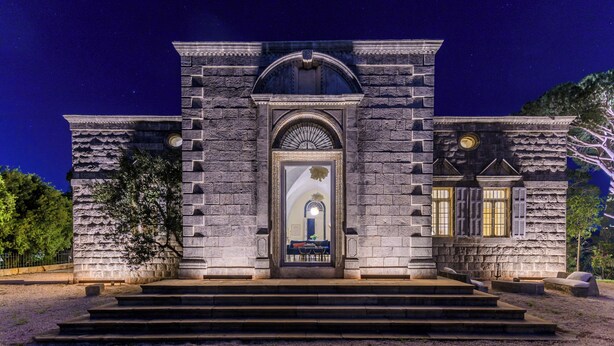 Un bâtiment en pierre doté d’une entrée centrale en arc de cercle, d’un escalier menant à l’entrée et d’une petite table avec des chaises à l’extérieur.