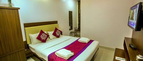 A hotel room with a bed, a wooden wardrobe, a desk with a chair, an air conditioning unit, and a television.