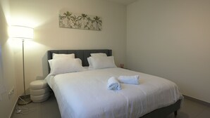 Una cama bien hecha con ropa de cama y almohadas blancas, una lámpara de noche y una obra de arte en la pared con palmeras.