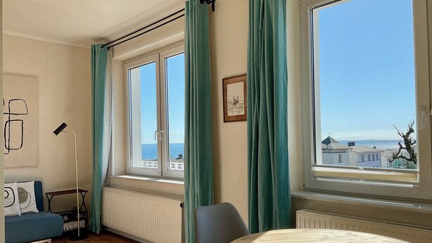 A room with a large window, a round table, a sofa, a radiator, and a framed picture on the wall.