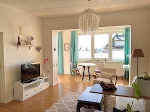 Ein Wohnzimmer mit Fernseher, einer Kristallleuchtre, einem Essbereich mit Stühlen, einem Tisch und einer Pflanze.