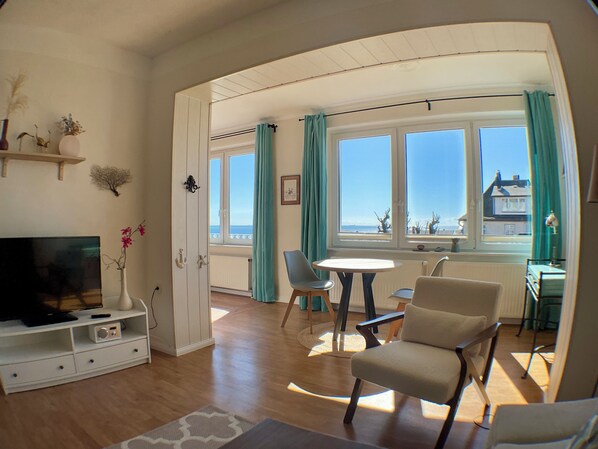 Ein Zimmer mit Fernseher, Esstisch, Stühlen, einer Couch und Meerblick durch große Fenster.
