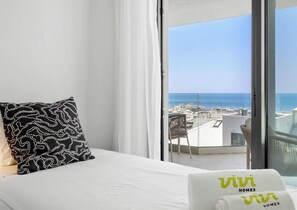 A hotel room with a bed, a patterned pillow, a white towel with 'vivi homes' branding, and a balcony with a view of the sea and buildings.