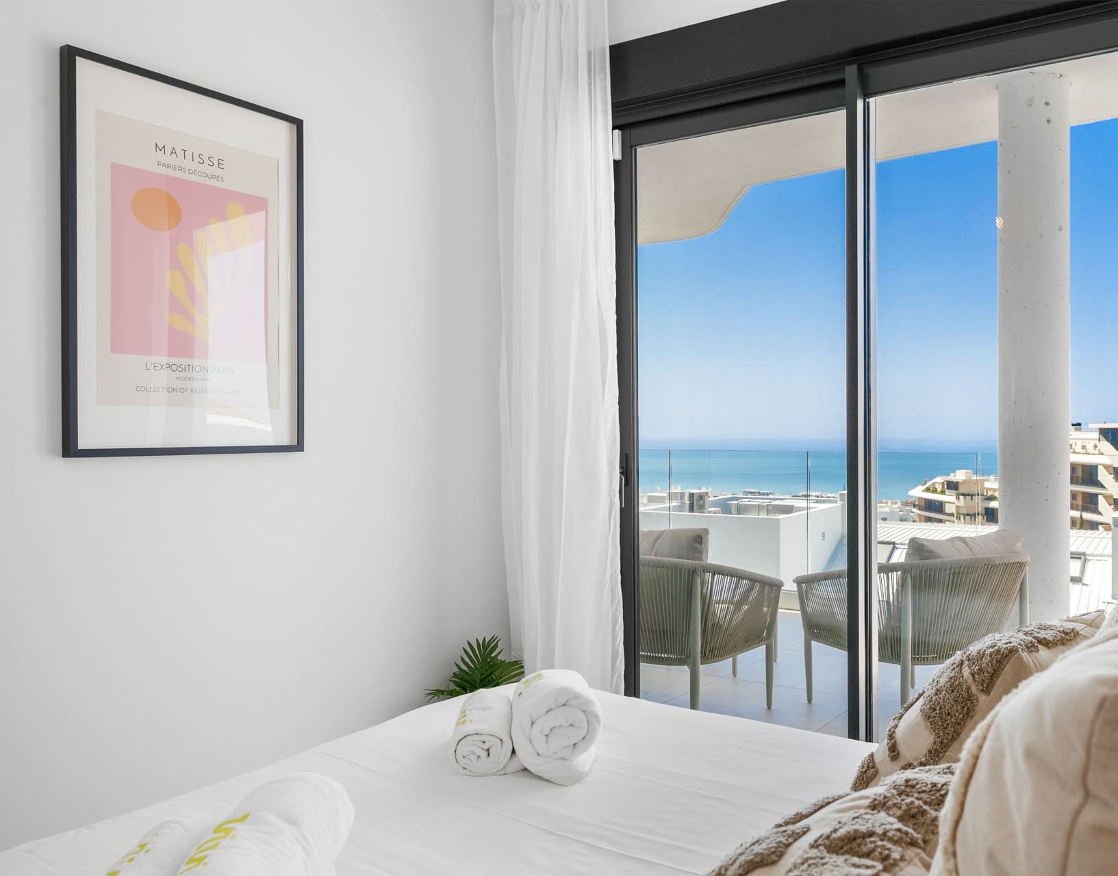 A hotel room with a bed, a framed artwork on the wall, a balcony with a view of the sea, and a logo in the corner.