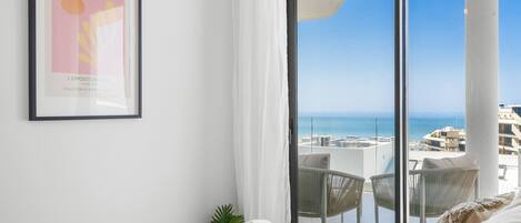 A hotel room with a bed, a framed artwork on the wall, a balcony with a view of the sea, and a logo in the corner.