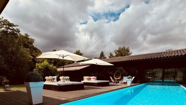 A swimming pool with lounge chairs and umbrellas, surrounded by a wooden deck and a building with a tiled roof. - Villa des chateaux du pessac leognan (Martillac)