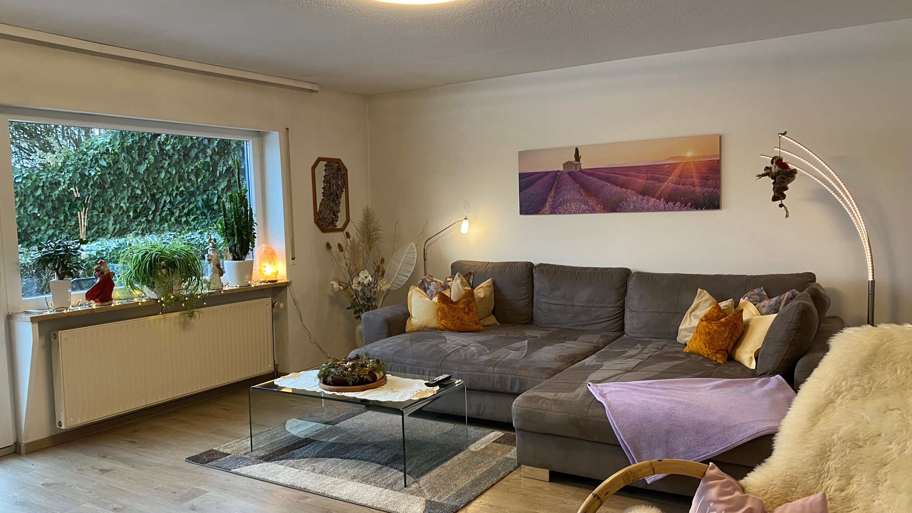 A living room with a grey sofa, a coffee table, a large window with plants, a painting, and a floor lamp.