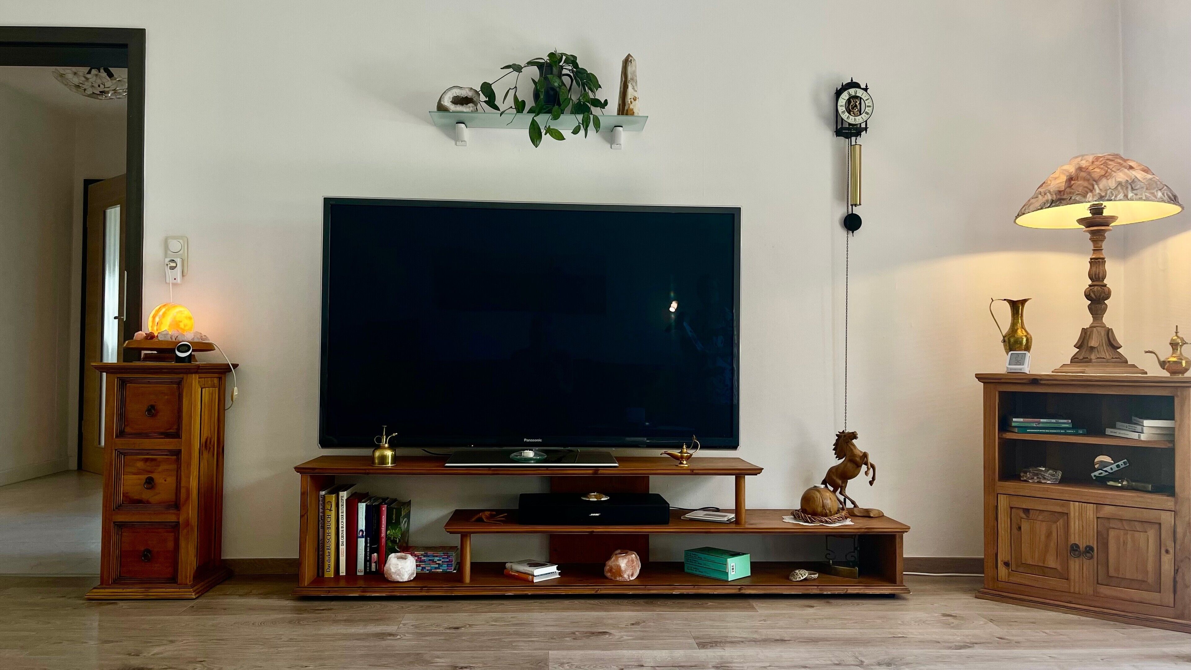 A living room with a large flat-screen TV, wooden entertainment unit, books, decorative items, and a wall-mounted clock.
