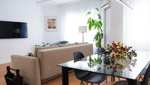 Een moderne woonkamer met een bank, een glazen eettafel en stoelen. Er is een tv aan de muur bevestigd, een plant en een lamp.