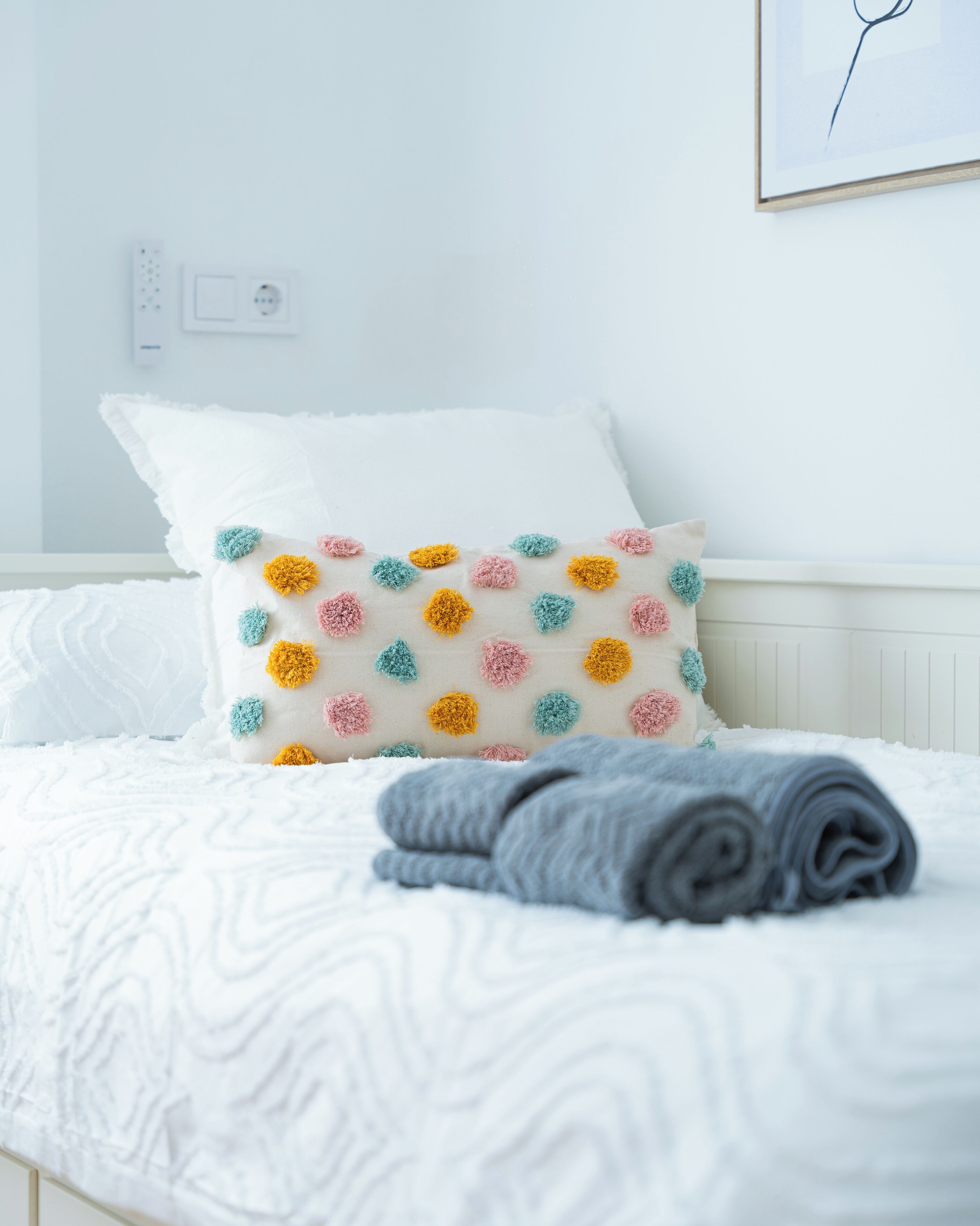 Ein ordentlich bezogenes Bett mit weißer Bettwäsche, ein farbenfrohes Kissen mit Pompons und eine aufgerollte graue Decke.