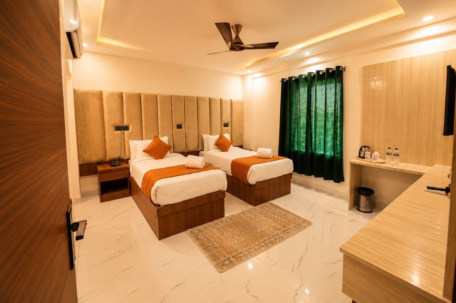 A modern hotel room with two beds, a wooden headboard, a green curtain, a small desk with toiletries, and a ceiling fan.