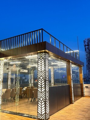 A modern outdoor seating area with glass partitions, wooden flooring, and a metal railing. - Hotel Dev Vilas Agra (Agra)