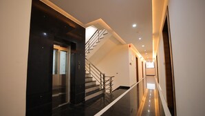 A modern hotel corridor with a polished black floor, a staircase with metal railings, and a glass elevator.