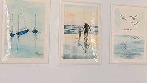 Three green glass bottles on a shelf above framed artworks depicting sailboats, a couple by the sea, and seagulls.