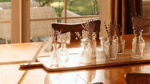 A dining table with chairs, a glass vase with dried flowers, and a view through a window with curtains.