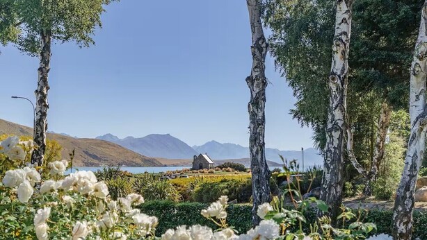 A serene landscape with a small stone building, surrounded by birch trees and white flowers, overlooking a body of water and distant mountains.