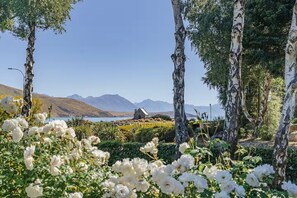 Un paisaje tranquilo con un pequeño edificio de piedra, rodeado de abedules y flores blancas, con vista a un cuerpo de agua y montañas distantes.