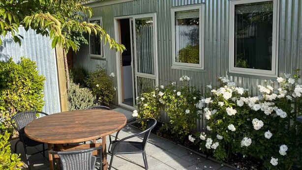 A metal-framed house with a wooden table and chairs outside, surrounded by white flowers and greenery. - Awa Cottage - Lake Tekapo (Lake Tekapo)