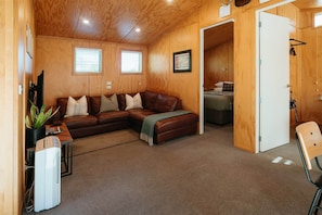 A cozy room with a brown leather sectional sofa, a small table with a plant, a white air conditioner, and wooden walls.