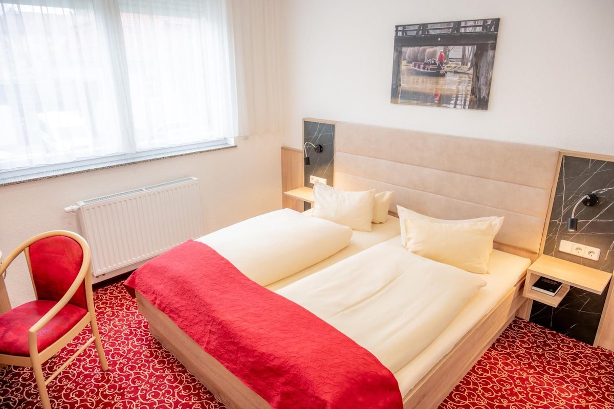 Ein Hotelzimmer mit einem Bett, einem Sessel, einem Nachttisch, einer Wandlampe und einem Fenster mit durchsichtigen Vorhängen.