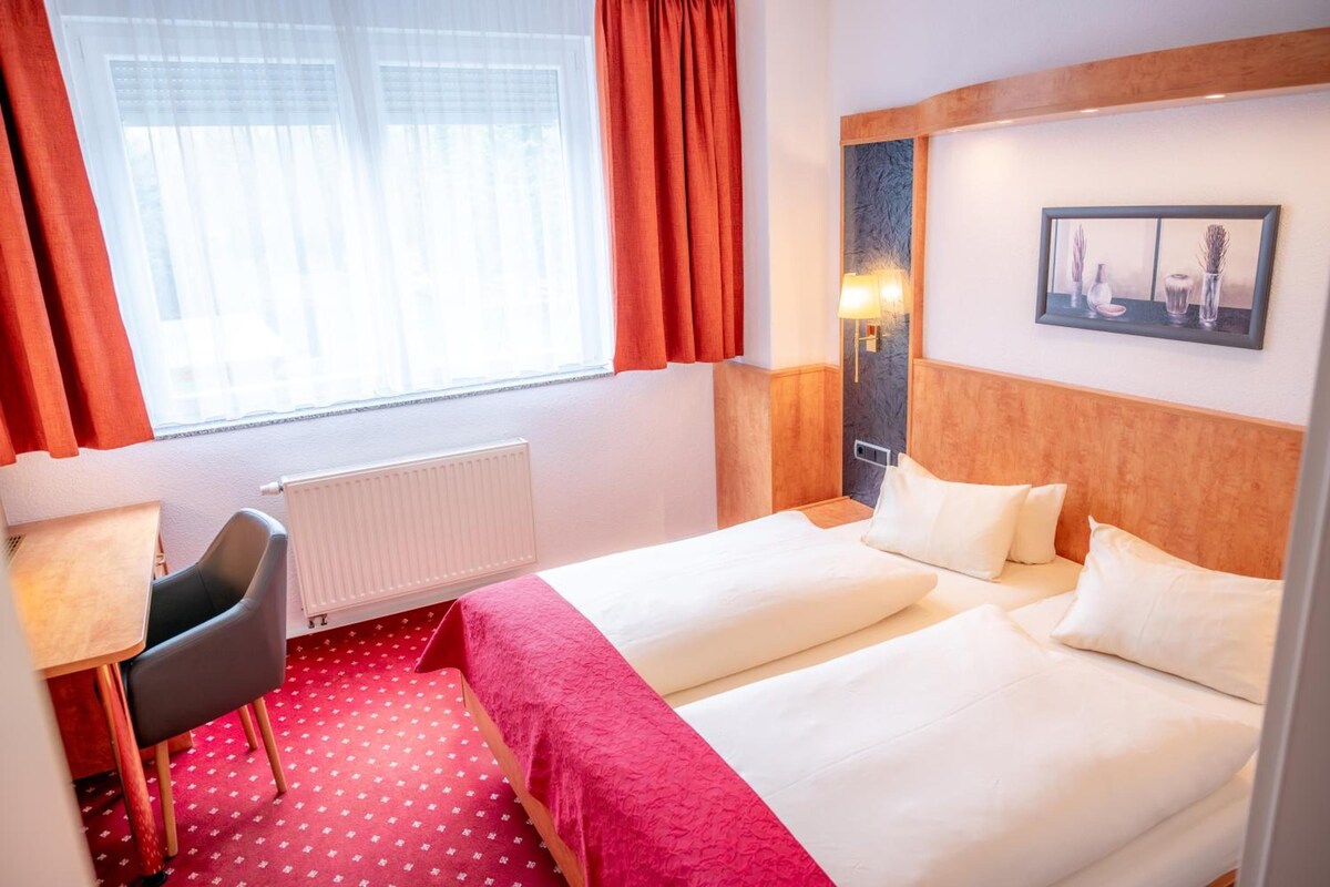 A hotel room with a bed, a desk, a chair, a radiator, and a window with curtains.