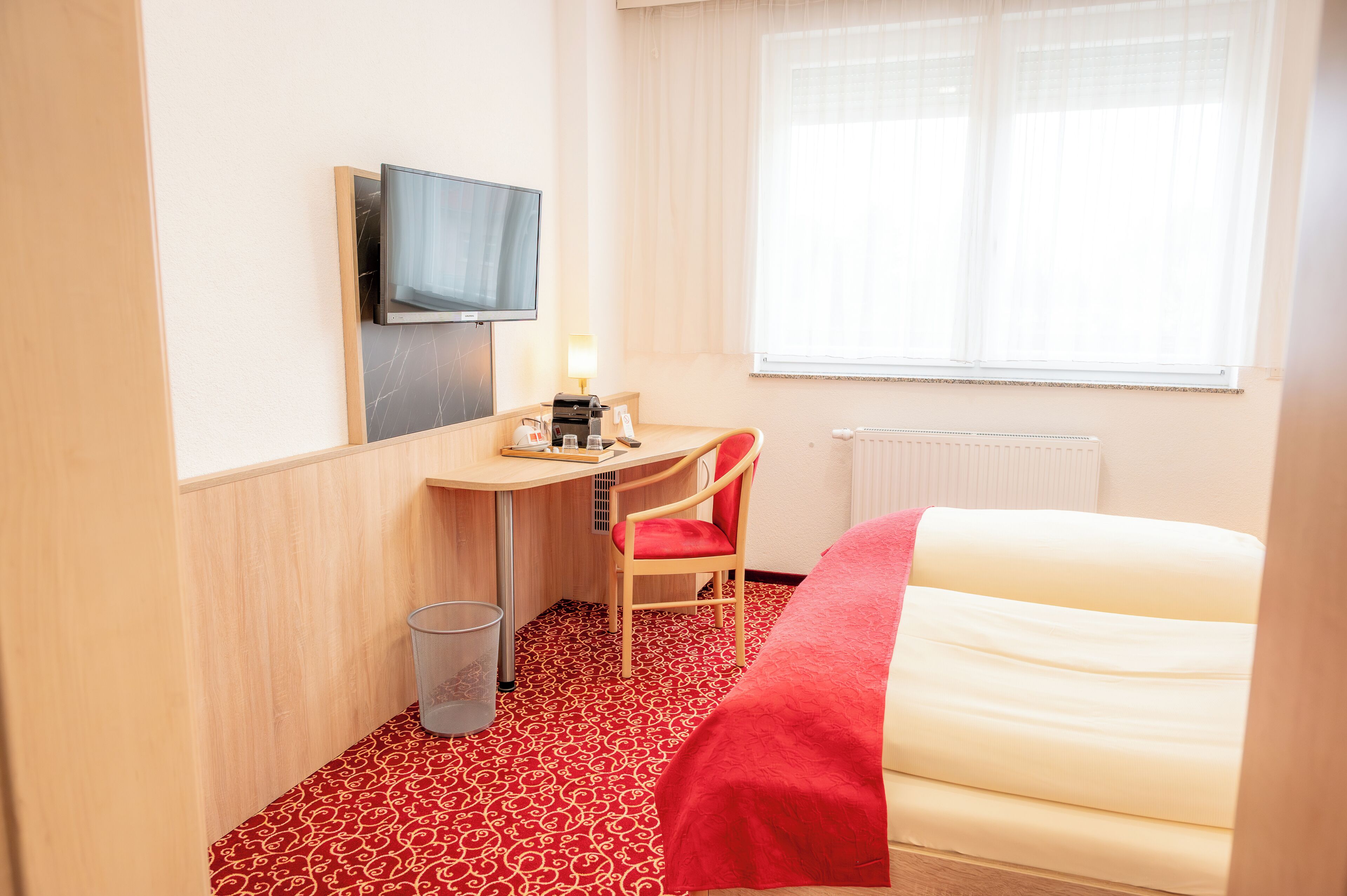 Una camera d'albergo con un letto, una scrivania con una sedia, un televisore a parete e una finestra con tende.