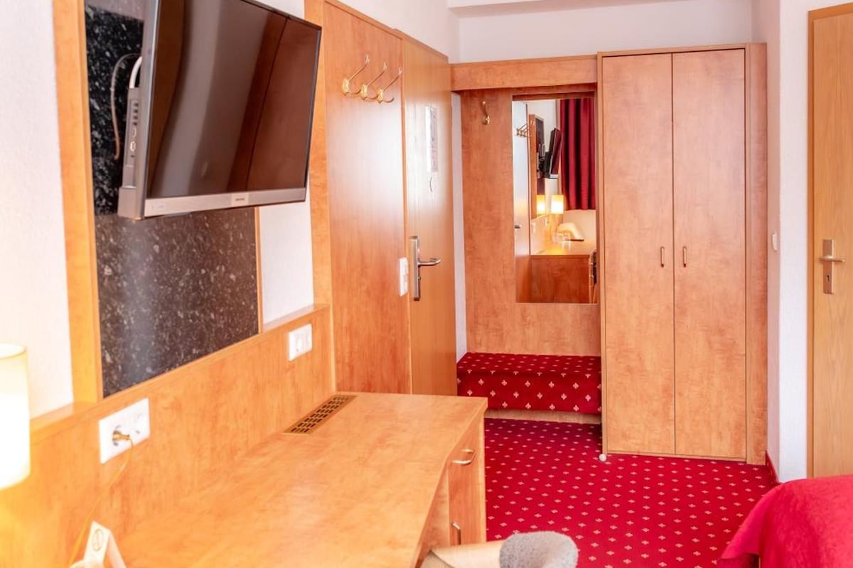 A hotel room with a wooden desk, a flat-screen TV, a bed with a red cover, and a wardrobe.