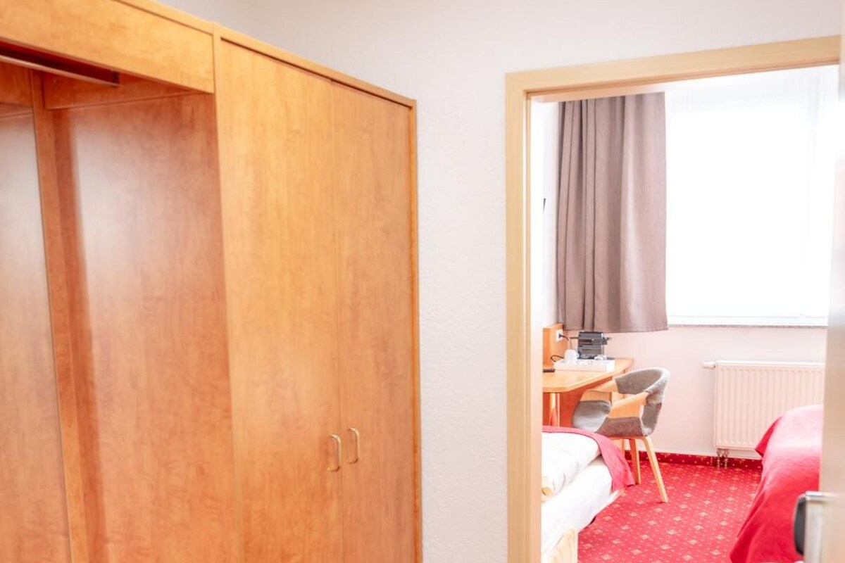 Une chambre d’hôtel équipée d’une armoire, d’un bureau avec une chaise et d’un lit recouvert d’une moquette rouge.