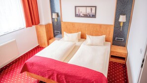 A double bed with a red blanket, two bedside tables with lamps, a framed picture above the bed, and a red carpeted floor. - AaRa Hotel (Radeberg)