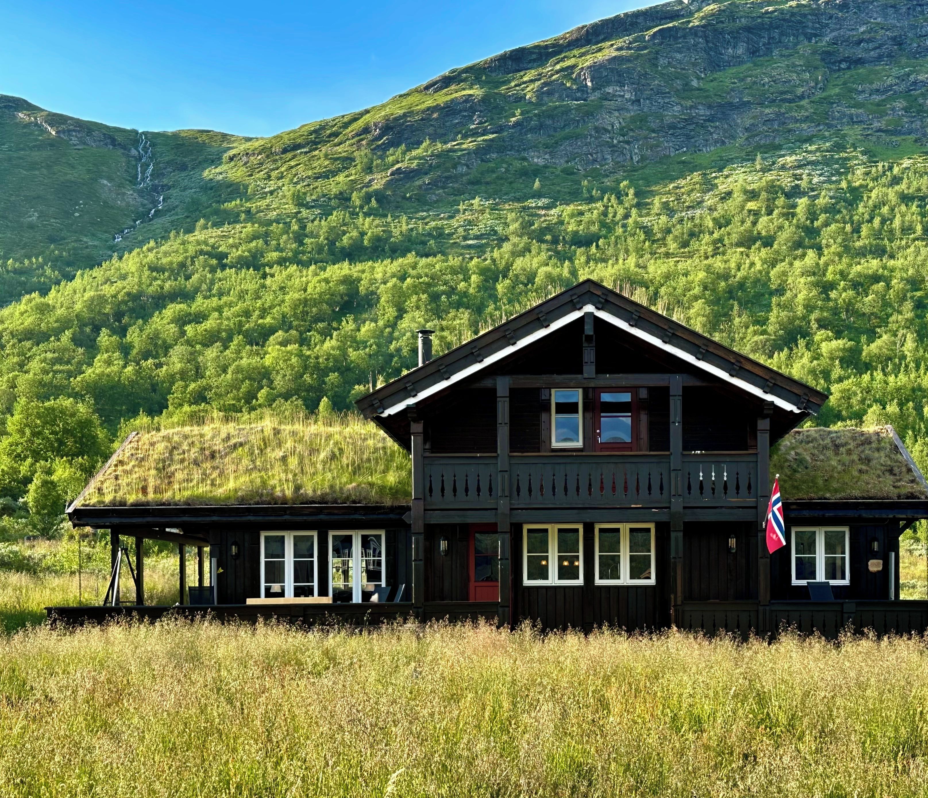 A wooden cabin with a thatched roof, surrounded by lush greenery and a mountainous backdrop.