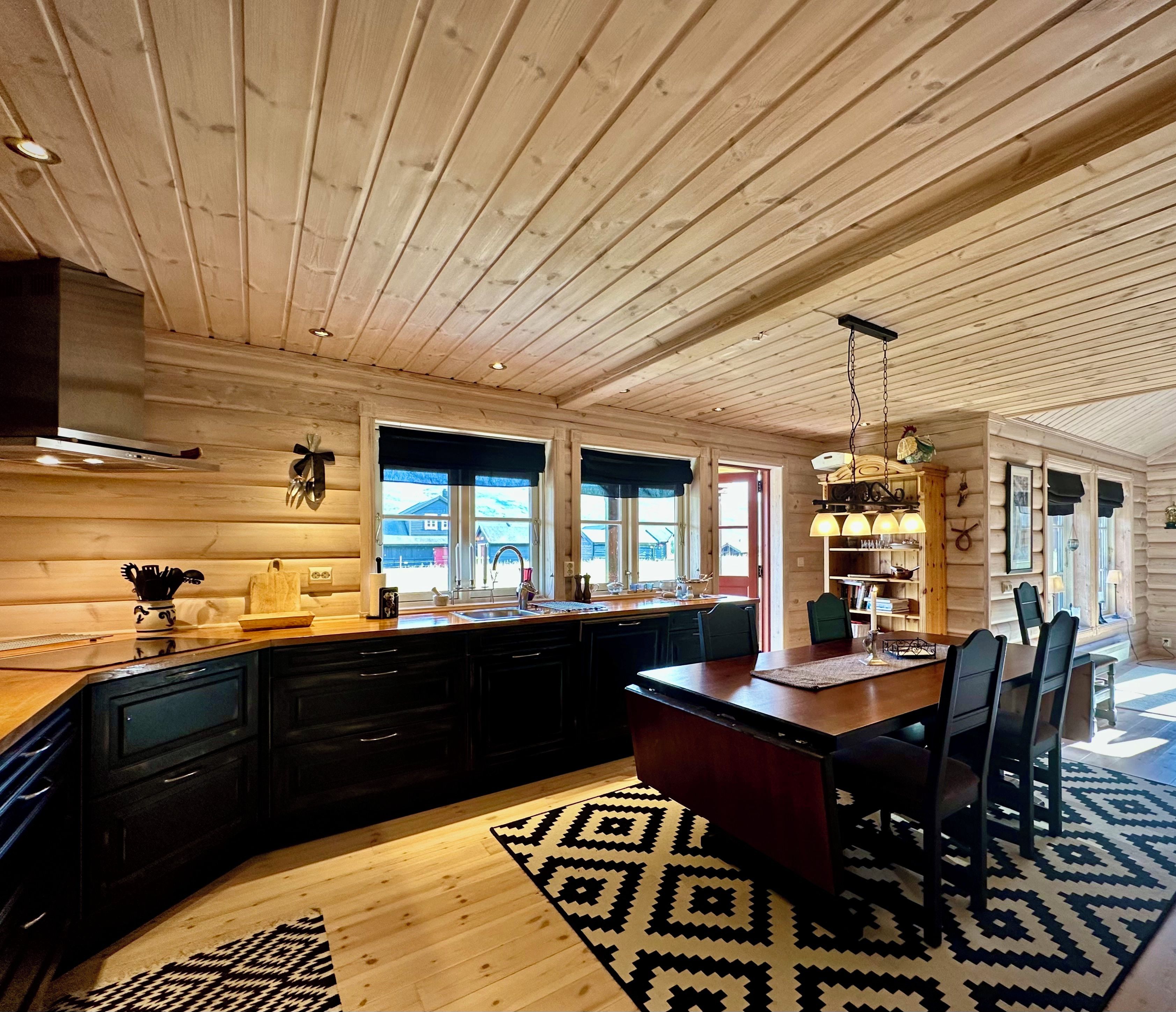 A log cabin interior with a kitchen and dining area, wooden ceiling, and large windows.