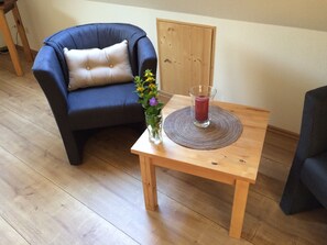 A room with a wooden floor, a dark blue armchair with a beige cushion, a small wooden table with a glass vase of flowers and a glass of red drink, and a wooden door in the background.