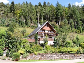 Uma casa em estilo chalé com varanda, cercada por um muro de pedra e vegetação exuberante.