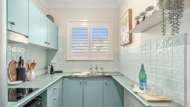 Een keuken met blauwgroene kastjes, een raam met jaloezieën, een spoelbak en een aanrecht met een fles en glazen.