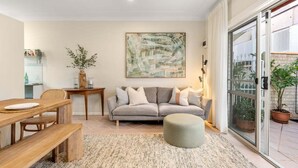 Een moderne woonkamer met een bank, een eettafel met stoelen, een salontafel, een vloerkleed, een schilderij aan de muur en een schuifpui naar een buitenruimte met planten.