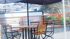 A balcony with outdoor seating, including tables and chairs, overlooking a waterfront area with buildings and trees. - Home of Comfort (Narok County)