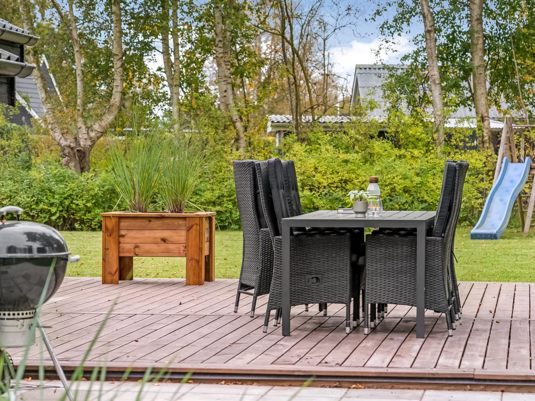 Um deck externo com mesa e cadeiras, churrasqueira, uma pequena mesa de madeira com plantas e um escorrega ao fundo.