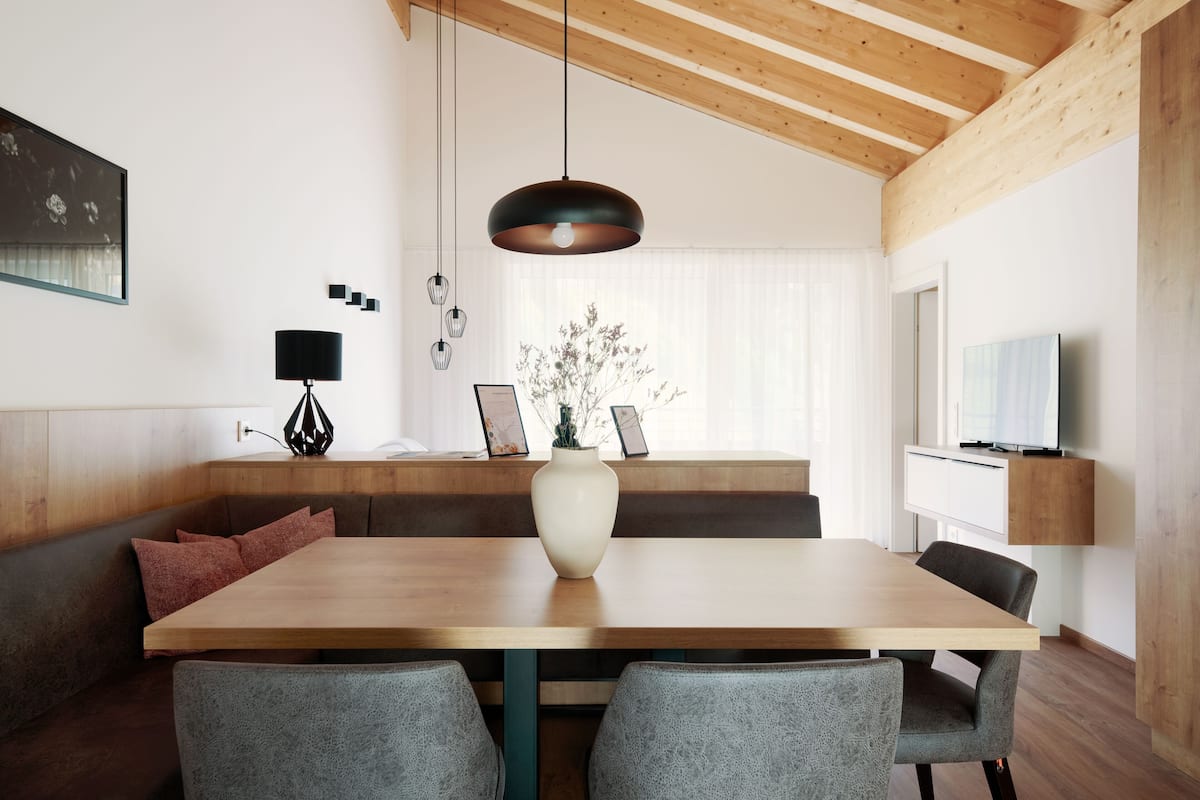A modern dining area with a wooden table, grey upholstered chairs, a white vase with flowers, and a pendant light.