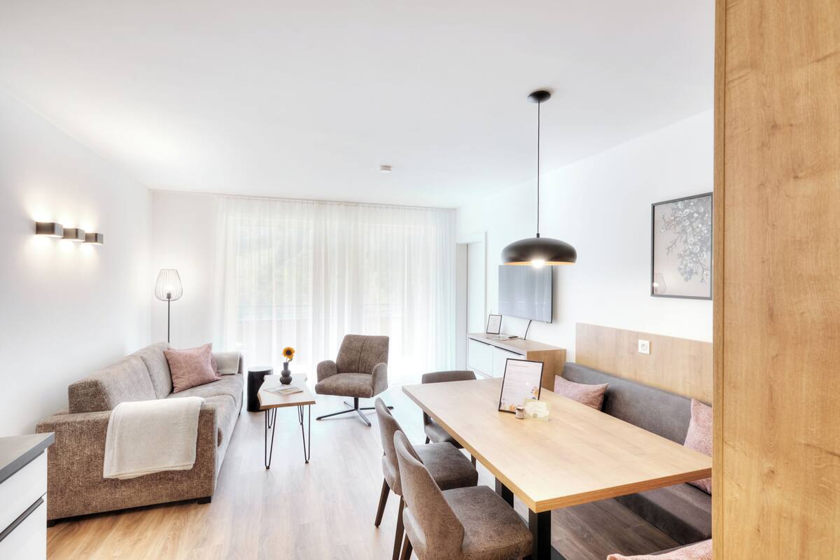A modern living room with a sofa, a dining table, and a chair. There is a window with curtains, a lamp, and a framed picture on the wall.