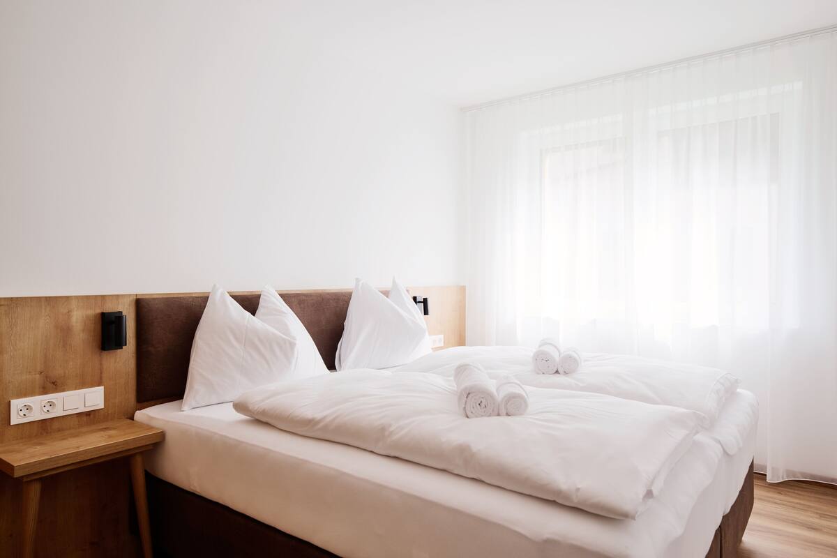 A hotel room with a neatly made bed, a wooden headboard, a bedside table with a power outlet, and a large window with sheer curtains.