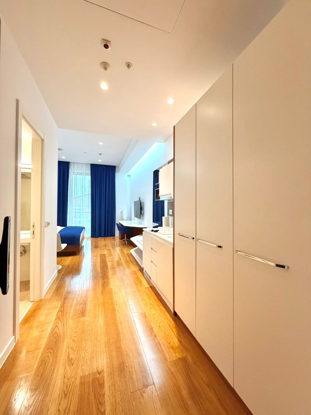 Ein modernes Hotelzimmer mit Holzboden, einem Bett mit blauen Vorhängen, einem Badezimmer mit Spiegel und einer Küchenzeile.